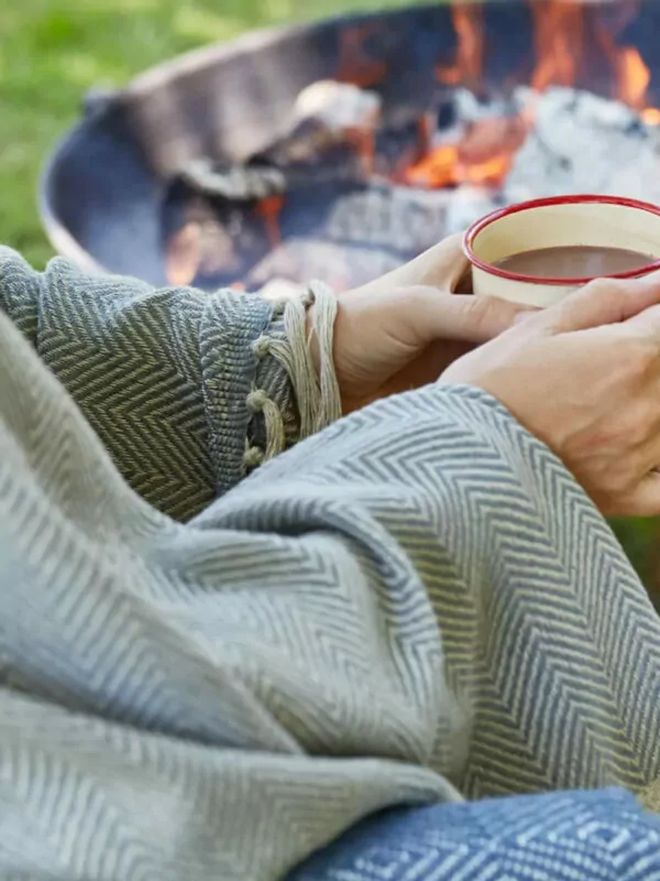Persoon buiten met warme kop koffie en deken om zich heen bij een open vuurtje