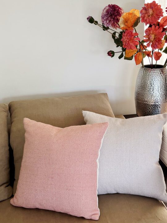 Koraal kleurig en heel licht roze gekleurd kussen op een bank met een boeket bloemen ernaast